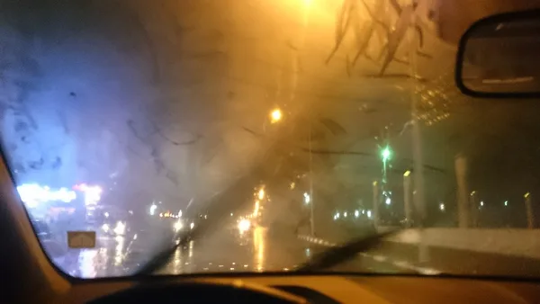 Foto dari kursi pengemudi MPV saat hujan deras, kaca depan berembun dan tangan pengemudi menekan tombol AC